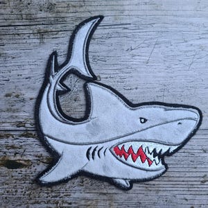 Può includere: Una toppa a forma di squalo grigio con contorno nero e bocca con denti rossi. La pinna dorsale dello squalo è sollevata e la toppa è per abbigliamento o accessori.