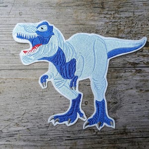 Könnte beinhalten: Gestickter Aufnäher eines Tyrannosaurus Rex Dinosauriers in Blau- und Weißtönen. Der Dinosaurier hat ein detailliertes Design mit sichtbaren Zähnen und Krallen. Der Aufnäher ist wahrscheinlich für Kleidung oder Accessoires.