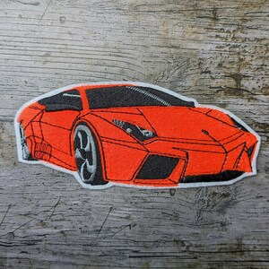 Pode incluir: Patch bordado com um carro esportivo laranja com detalhes em preto e uma borda branca. O design do carro inclui faróis e rodas, em um fundo de madeira texturizada.