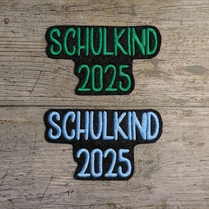 Könnte beinhalten: Zwei gestickte Aufnäher auf einer Holzoberfläche. Der obere Aufnäher hat grünen Text mit der Aufschrift "SCHULKIND 2025". Der untere Aufnäher hat blauen Text mit der Aufschrift "SCHULKIND 2025". Beide Aufnäher sind schwarz.