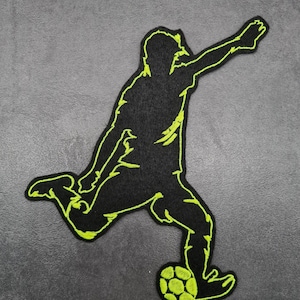 May include: Black and neon green embroidered patch featuring a soccer player kicking a ball. The patch depicts a silhouette of a soccer player in action, with the ball at their foot. The design is outlined in neon green.