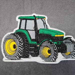 May include: Embroidered patch of a green tractor with black tires and yellow wheel centers. The tractor has a white border and is set against a gray background. The tractor's design includes a cab and detailed tire treads.