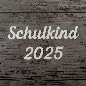 May include: White text on a weathered wood background reads "Schulkind 2025". The text is in a cursive font, with the numbers in a simple, bold font. The wood has a gray and brown color.