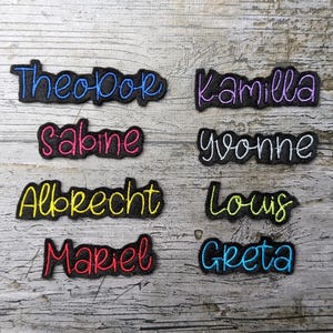 Peut inclure: Écussons brodés avec des noms de différentes couleurs, dont le bleu, le rose, le jaune et le rouge. Les noms incluent Theobok, Kamilla, Sabine, Yvonne, Albrecht, Louis, Mariel et Greeta. Chaque écusson a une bordure noire.