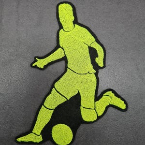 Könnte beinhalten: Ein neongrüner, gestickter Aufnäher mit einem Fußballspieler in Aktion, der einen Ball tritt. Der Spieler wird in einer dynamischen Pose mit schwarzer Umrandung dargestellt. Der Aufnäher ist für das Aufnähen auf Kleidung oder Accessoires konzipiert.