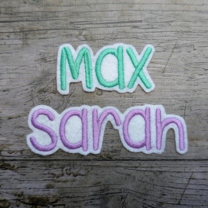 Peut inclure: Deux écussons brodés avec des noms. Le patch supérieur indique "MAX" en fil vert avec un contour blanc. Le patch inférieur indique "sarah" en fil violet avec un contour blanc. Les écussons sont sur un fond en bois.