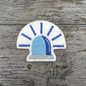 Op de afbeelding: Geborduurde patch met een blauw en wit noodsirene-ontwerp. De patch heeft een witte achtergrond met blauwe accenten en een lichtblauwe sirenevorm in het midden. De patch is ongeveer 5 cm breed.