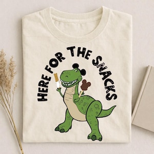 May include: Cream-colored t-shirt featuring a green cartoon dinosaur holding ice cream. The text "HERE FOR THE SNACKS" arches above the dinosaur. The dinosaur has a black hair-like design on its head and a light brown belly.