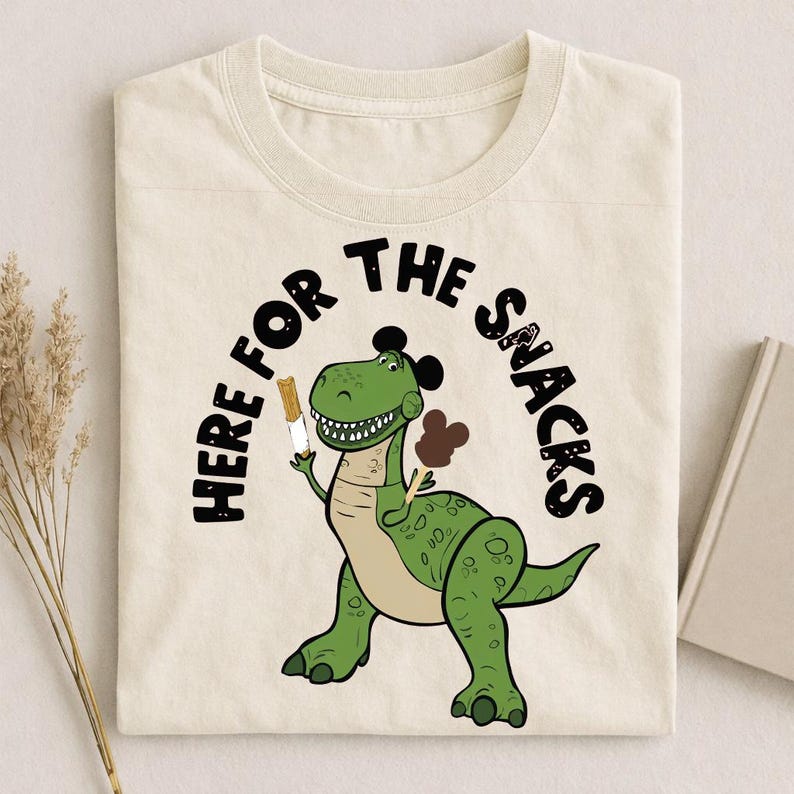 May include: Cream-colored t-shirt featuring a green cartoon dinosaur holding ice cream. The text "HERE FOR THE SNACKS" arches above the dinosaur. The dinosaur has a black hair-like design on its head and a light brown belly.