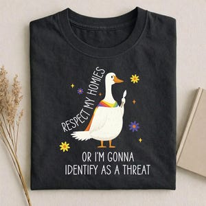 May include: Black t-shirt featuring a white goose with a rainbow-colored wing and a knife. The text reads "Respect My Homies" and "Or I'm Gonna Identify As A Threat". Yellow and purple floral accents surround the goose.
