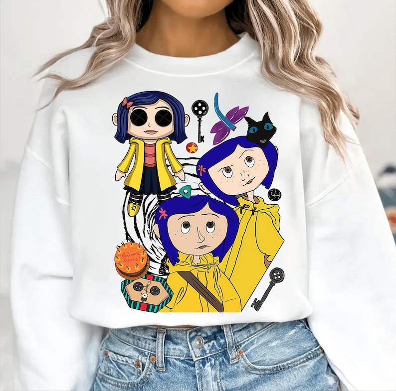May include: White sweatshirt featuring a colorful graphic of a cartoon character in a yellow raincoat. The design includes multiple illustrations of the character, keys, and a black cat. The sweatshirt is a casual, comfortable style.