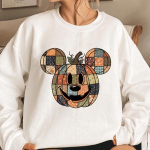 May include: White sweatshirt with a pumpkin design incorporating Mickey Mouse ears. The pumpkin is crafted from patchwork fabric in orange, blue, and green floral patterns. The design features a smiling face and a stem with leaves.