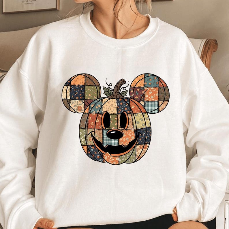 May include: White sweatshirt with a pumpkin design incorporating Mickey Mouse ears. The pumpkin is crafted from patchwork fabric in orange, blue, and green floral patterns. The design features a smiling face and a stem with leaves.