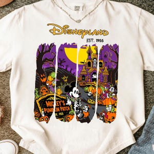 Puede incluir: Camiseta blanca con un diseño de Halloween de Disneyland. El diseño presenta a Mickey Mouse con un disfraz de esqueleto, calabazas y una casa embrujada. También se ve el texto "Disneyland Est. 1955" y "Mickey's Pumpkin Patch Est. 1928".