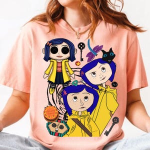 May include: A light pink t-shirt featuring a graphic design of a cartoon character with blue hair and a yellow raincoat. The design includes a black cat, keys, and other elements from the movie.
