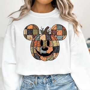 May include: White sweatshirt with a patchwork pumpkin design incorporating Mickey Mouse ears. The pumpkin is in shades of orange, green, and blue, with a smiling face and a stem. The design is centered on the front of the shirt.