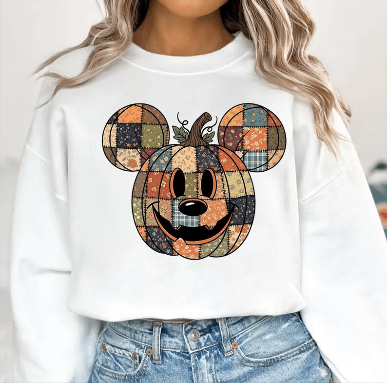 May include: White sweatshirt with a patchwork pumpkin design incorporating Mickey Mouse ears. The pumpkin is in shades of orange, green, and blue, with a smiling face and a stem. The design is centered on the front of the shirt.