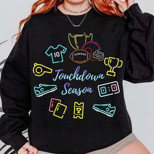 Puede incluir: Sudadera negra con ilustraciones de neón coloridas de artículos relacionados con el fútbol. El diseño incluye un balón de fútbol, casco, trofeo, zapatillas y las palabras "Touchdown Season". El número 10 está en una camiseta.
