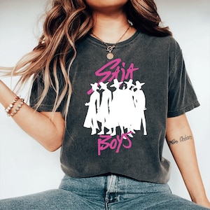 Op de afbeelding: Donkergrijs t-shirt met de roze tekst "Sia Boys" en een wit silhouet van figuren met hoeden. Het t-shirt heeft een vintage, versleten uitstraling.