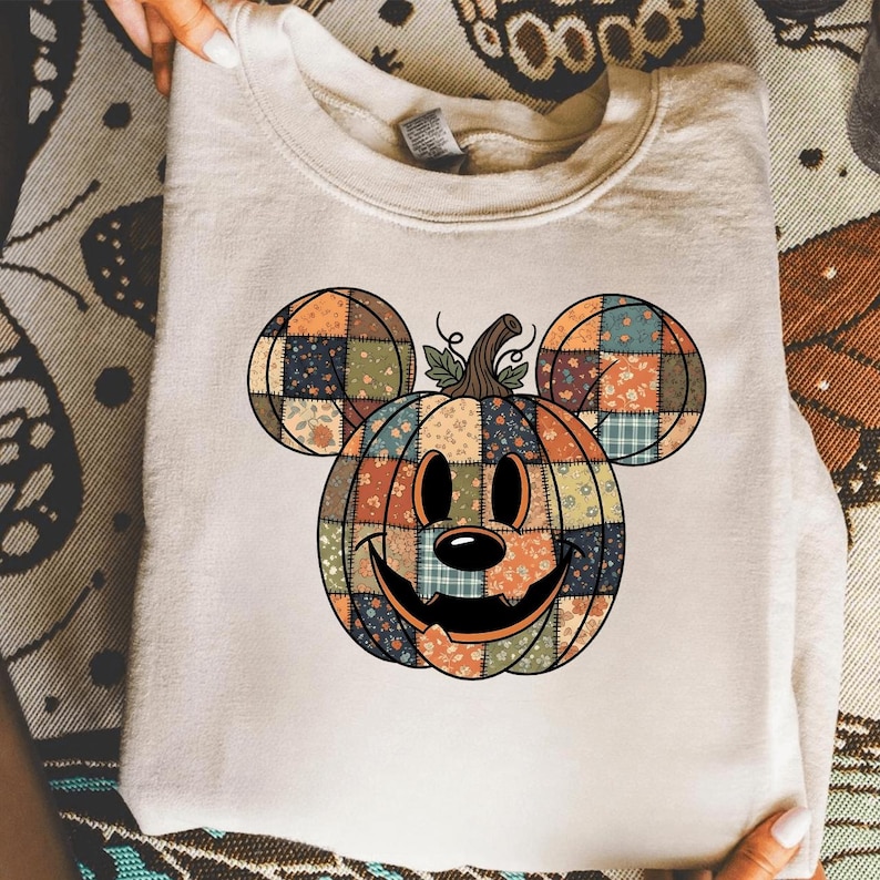 May include: A cream-coloured sweatshirt with a colourful Mickey Mouse-shaped pumpkin design. The pumpkin is made of patchwork squares in shades of orange, green, and brown, with a smiling face and stem. The ears are also patchwork.