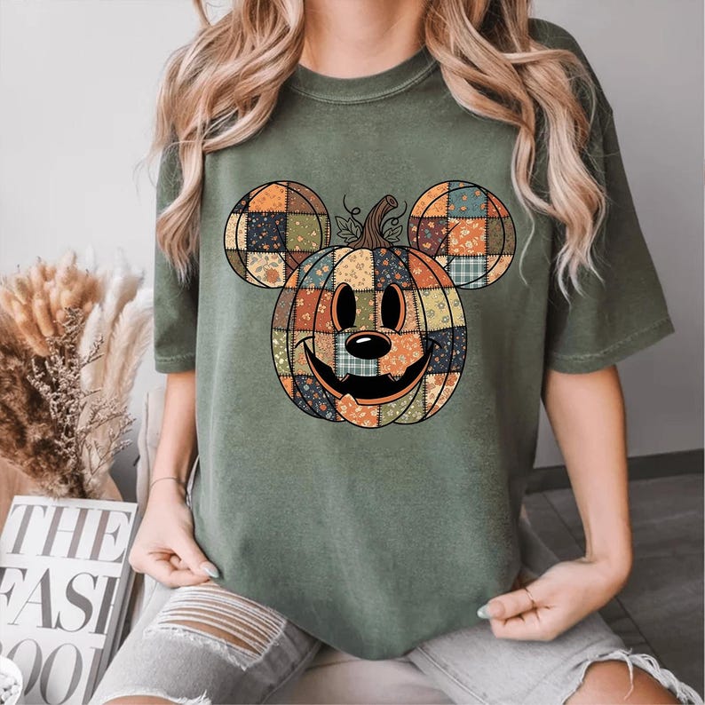 May include: An olive green t-shirt with a patchwork pumpkin design incorporating Mickey Mouse ears. The pumpkin has a smiling face and is made of various patterned fabrics in orange, blue, and yellow.