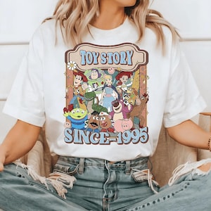 Può includere: T-shirt bianca con grafica retrò dei personaggi di Toy Story. Il design include le parole "TOY STORY" e "SINCE 1995" in marrone e blu. La maglietta presenta vari personaggi del film d'animazione, in stile vintage.