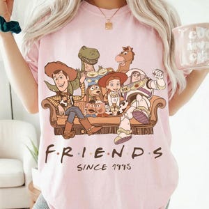 May include: A light pink t-shirt featuring a graphic of characters from the Toy Story movie franchise. The word "FRIENDS" is printed in a stylized font, with "SINCE 1995" below. The design includes Woody, Buzz Lightyear, and other characters.