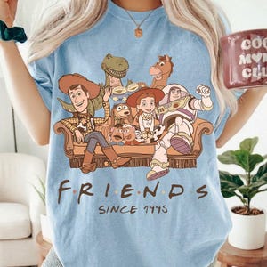 May include: Light blue t-shirt featuring a graphic of Toy Story characters on a brown couch. The word "FRIENDS" is printed below the graphic, with "SINCE 1995" underneath. The shirt is a light blue color.