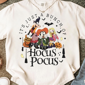 May include: Cream-colored t-shirt with a graphic featuring cartoon characters and the text "IT'S JUST A BUNCH OF HOCUS POCUS." The design includes pumpkins, bats, and ghosts, perfect for Halloween.