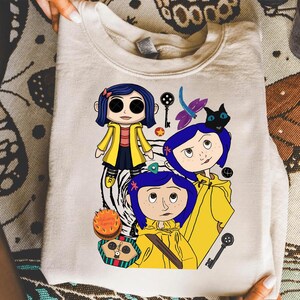 May include: Cream-colored sweatshirt featuring a graphic of a cartoon character with blue hair and a yellow raincoat. The design includes a black cat, keys, and a dragonfly. The sweatshirt is folded, with the design prominently displayed.