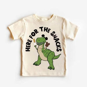 May include: Cream-colored t-shirt featuring a cartoon dinosaur holding a snack. The dinosaur is green with brown accents and a black outline. The text "HERE FOR THE SNACKS" arches above the dinosaur in black letters.