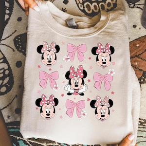 Puede incluir: Sudadera de color crema con un patrón repetido de caras de Minnie Mouse, lazos rosas con lunares y lazos rosas. El diseño incluye pequeñas estrellas rosas y flores blancas, creando una estética lúdica y femenina.