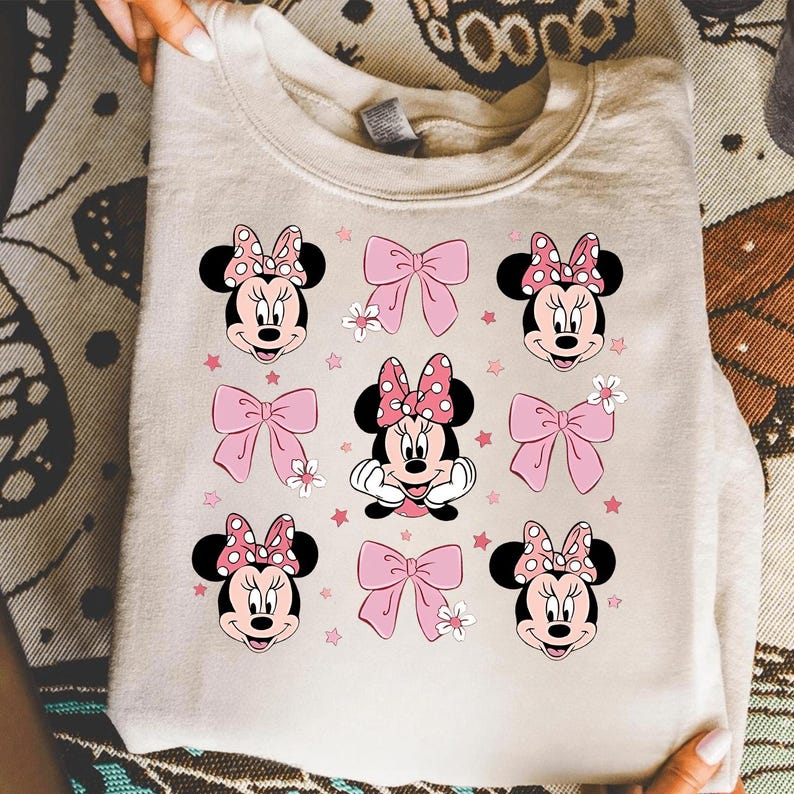 May include: Cream-colored sweatshirt featuring a repeating pattern of Minnie Mouse faces with pink polka-dot bows and pink bows. The design includes small pink stars and white flowers, creating a playful and feminine aesthetic.