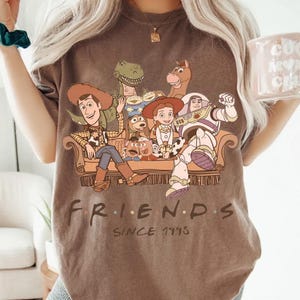 May include: Brown t-shirt featuring a graphic of Toy Story characters sitting on a couch. The word "FRIENDS" is printed below the image, with "SINCE 1995" underneath. The shirt has a vintage, worn look.