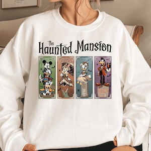 May include: White sweatshirt featuring a graphic from The Haunted Mansion. The design includes Mickey Mouse, Minnie Mouse, Donald Duck, and Goofy in spooky attire. The text "The Haunted Mansion" is printed above the characters.