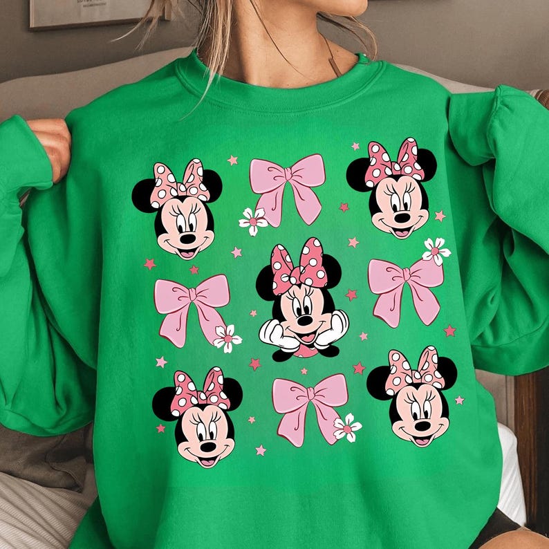 May include: Green sweatshirt featuring a Minnie Mouse design. The design includes multiple Minnie Mouse faces with pink polka dot bows, pink bows, white flowers, and pink stars. The sweatshirt is a solid green color.
