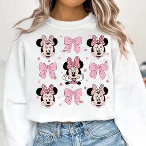 May include: White sweatshirt featuring a repeating pattern of Minnie Mouse faces with pink polka dot bows, pink bows, and pink stars. The design is centered on the front of the sweatshirt.