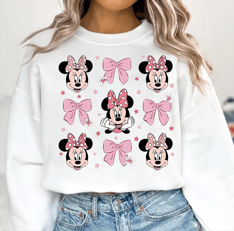 May include: White sweatshirt featuring a repeating pattern of Minnie Mouse faces with pink polka dot bows, pink bows, and pink stars. The design is centered on the front of the sweatshirt.
