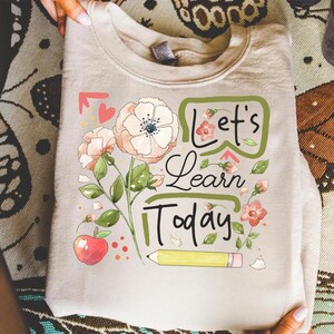 May include: A light beige sweatshirt featuring a floral design with the words "Let's Learn Today." The design includes watercolor-style flowers, an apple, and a pencil. The sweatshirt is made of a soft material.