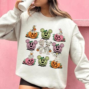 May include: A light beige sweatshirt featuring a Halloween-themed design. The design includes cartoon characters dressed as mummies and pumpkins with Mickey Mouse ears. The pumpkins are orange, green, and pink.
