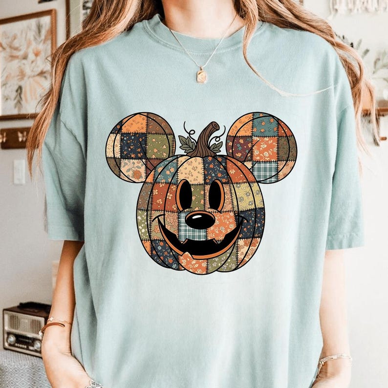 May include: A pale blue t-shirt with a patchwork pumpkin design incorporating Mickey Mouse ears. The pumpkin is made of various fabric patterns in orange, brown, and green. The design has a smiling face, ideal for Halloween.