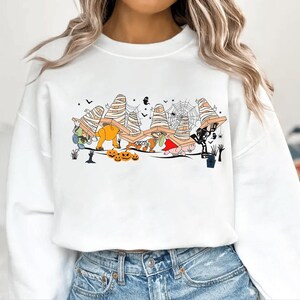 Puede incluir: Sudadera blanca con un diseño de Halloween. El diseño presenta personajes de dibujos animados disfrazados de momias, calabazas y otras figuras espeluznantes. Murciélagos negros, telarañas y una lápida completan la escena festiva.
