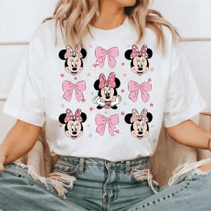 May include: White t-shirt featuring a repeating pattern of Minnie Mouse faces, pink bows, and stars. The Minnie Mouse faces have black ears and pink polka dot bows. The bows are a solid pink color. The shirt is a casual style.