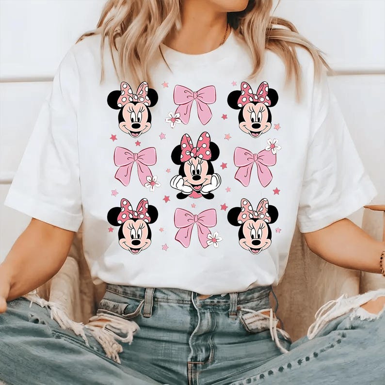 May include: White t-shirt featuring a repeating pattern of Minnie Mouse faces, pink bows, and stars. The Minnie Mouse faces have black ears and pink polka dot bows. The bows are a solid pink color. The shirt is a casual style.