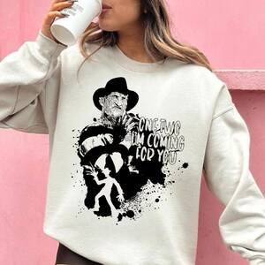 以下が含まれることがあります： クリーム色のスウェットシャツ。黒と白のグラフィックで、帽子とストライプのセーターを着たホラー映画のキャラクターが描かれています。「ONE TWO I'M COMING FOR YOU」の文字が前面にプリントされています。デザインにはスプラッター効果が含まれています。