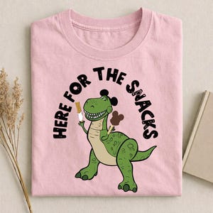 May include: Pink t-shirt featuring a green dinosaur holding a snack, with the text "HERE FOR THE SNACKS" in a black arch. The dinosaur has a light brown belly and black accents. The shirt is made of a soft material.