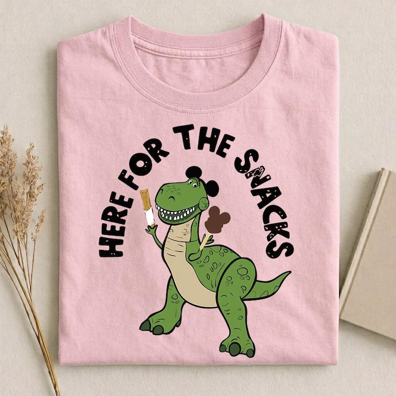May include: Pink t-shirt featuring a green dinosaur holding a snack, with the text "HERE FOR THE SNACKS" in a black arch. The dinosaur has a light brown belly and black accents. The shirt is made of a soft material.