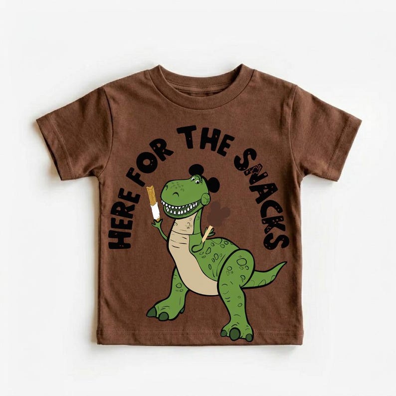 May include: Brown t-shirt featuring a green cartoon dinosaur holding a snack. The text "HERE FOR THE SNACKS" arches above the dinosaur. The dinosaur has a light tan belly and black accents.