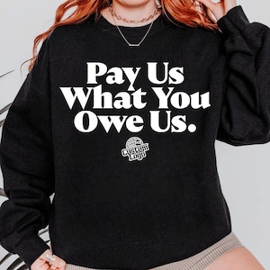 Può includere: Una felpa nera con la scritta bianca "Pay Us What You Owe Us" a caratteri grandi. Sotto il testo c'è un piccolo logo che dice "Custom Logo" con un design di basket. La felpa è in stile casual.