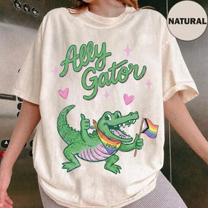 Puede incluir: Camiseta color crema con el texto "Ally Gator" en letras verdes y detalles rosas. Un caimán verde con una bandera y pañuelo Pride arcoíris es el gráfico central. La camiseta tiene mangas cortas.